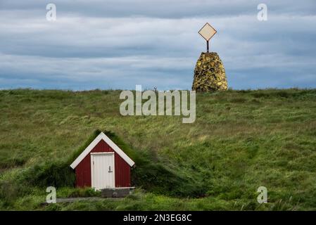 Structure de stockage sur le côté d'une colline herbeuse et formation de pierre avec réflecteur radar sur le sommet de la colline sur l'île Flatey, un ancien village de pêcheurs... Banque D'Images