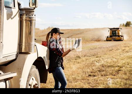 Une femme debout à côté d'un transporteur de grains, utilisant des appareils portables sans fil pour gérer et surveiller une récolte de canola d'automne, parlant au téléphone, avec un c... Banque D'Images