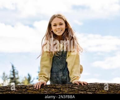 Jeune fille jouant sur une branche d'arbre et posant pour une photo dans un parc de la ville par un chaud après-midi d'automne ; tenue Albert, Alberta, Canada Banque D'Images