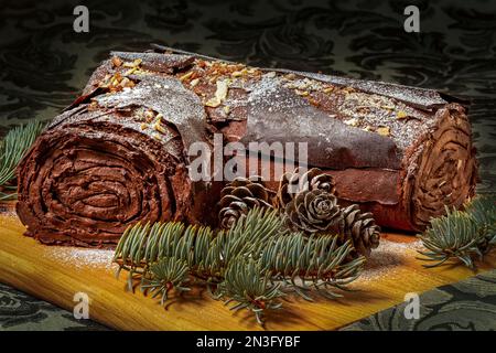 Glaçage roulé au chocolat glacé gâteau de bois de yule avec des morceaux d'arbre à feuilles persistantes et des cônes de pin sur une planche en bois assis sur une table décorative vert foncé c... Banque D'Images