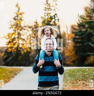 Un père portant sa jeune fille sur ses épaules tout en marchant dans un parc de la ville pendant la saison d'automne; St. Albert, Alberta, Canada Banque D'Images