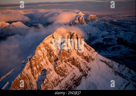 Vue aérienne sur le Rocky Mountain Front du Montana, avec des sommets enneigés des montagnes Rocheuses illuminés par la lumière du coucher du soleil au-dessus des nuages Banque D'Images