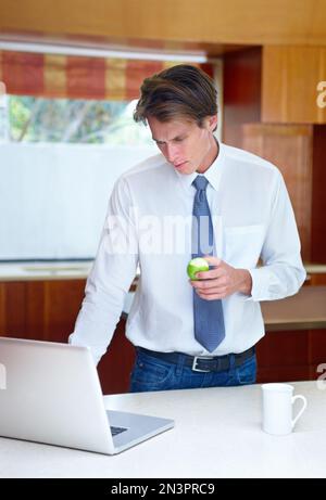 La première chose à faire pour recevoir des e-mails. Un jeune homme qui navigue sur son ordinateur portable dans la cuisine tout en dégustant un en-cas. Banque D'Images