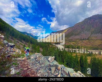 Fille voyageur avec un sac à dos sur des bâtons de randonnée grimpant sur le chemin de montagne escarpé en pierre à Altai avec la toile de fond d'une falaise et d'une rivière. Banque D'Images