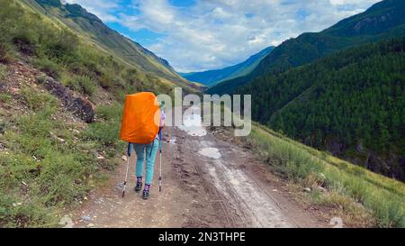 Une fille touristique avec un grand sac à dos orange et des bâtons de randonnée marche fatigués le long de la route en été près des montagnes et des nuages de l'Altaï. Banque D'Images