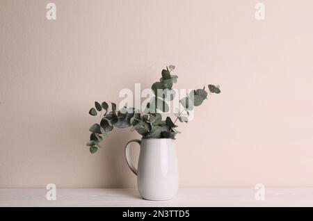 Vase avec de belles branches d'eucalyptus sur une table en bois blanc près d'un mur beige Banque D'Images