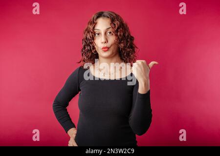 Portrait d'une jeune femme à tête rouge portant une robe à côtes noires isolée sur fond rouge points perplexes avec son pouce à l'espace de copie vide et le regard Banque D'Images