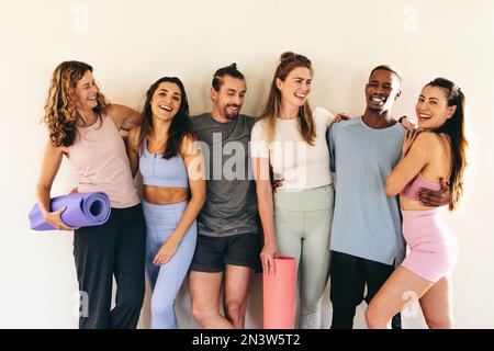 Diverses personnes rient avec joie tout en se tenant contre un mur avec des tapis de yoga. Personnes multiculturelles assistant à un cours de yoga ensemble. Groupe de sport pe Banque D'Images