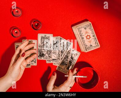 Composition magique ésotérique vue de dessus avec les mains de femme de la fortune teller avec la lecture de cigarettes cartes de Tarot sur fond rouge avec des bougies allumées Banque D'Images