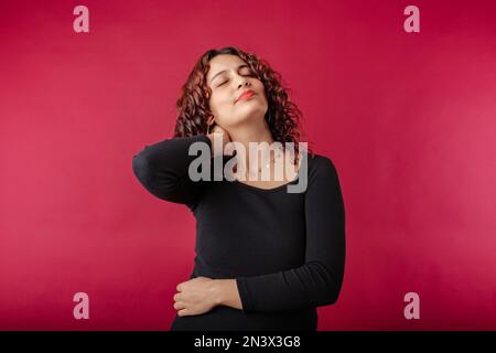 Portrait d'une jeune femme à tête rouge portant une robe à côtes noires isolée sur fond rouge avec une incroyable douleur au cou semble malheureux et des touches ou un massage Banque D'Images