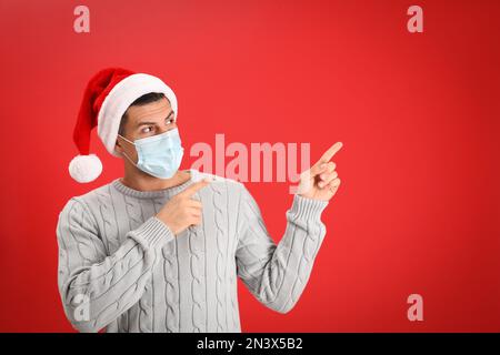 Homme portant un chapeau de père Noël et un masque médical sur fond rouge Banque D'Images
