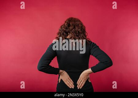 Jeune femme caucasienne portant une robe noire striée debout isolée sur fond rouge avec une douleur très sévère de dos retient avec la main et montre le p Banque D'Images