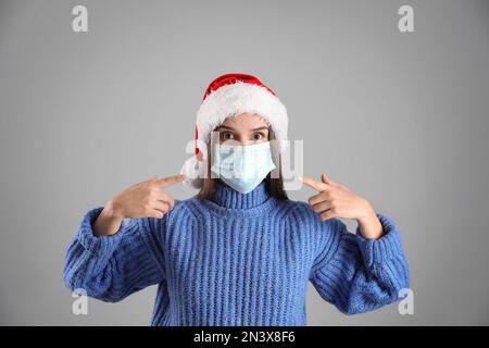 Femme confuse en chapeau de père Noël et masque médical sur fond gris Banque D'Images