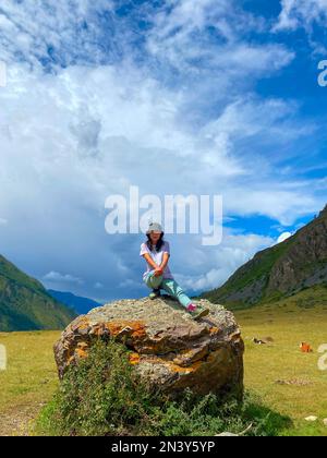 Une fille de voyageur asiatique dans un chapeau et des lunettes s'assoit avec sa jambe étirée sur un fond de pierre des montagnes de l'Altaï. Cadre vertical. Banque D'Images