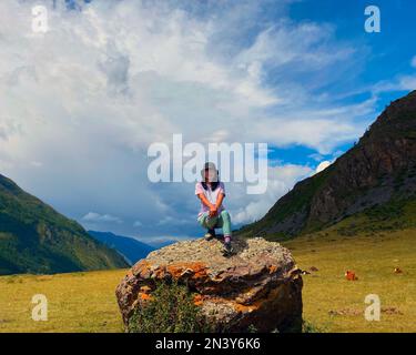 Une fille de voyageur asiatique dans un chapeau et des lunettes s'assoit avec sa jambe étirée sur un fond de pierre des montagnes dans la soirée Altaï. Banque D'Images