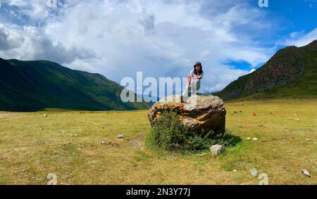 Une fille de voyageur asiatique dans un chapeau et des lunettes s'assoit avec sa jambe étirée sur un fond de pierre des montagnes de l'Altaï. Banque D'Images