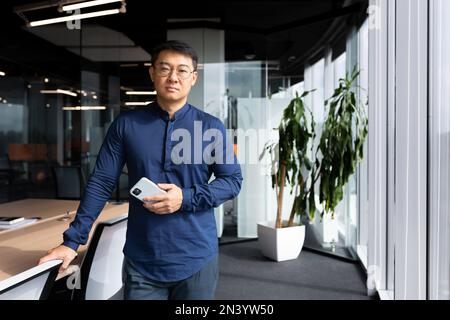 Portrait d'un homme asiatique sérieux à l'intérieur du bureau, d'un homme en lunettes et d'une chemise regardant la caméra, développeur de logiciels de programmation mature sur le lieu de travail tenant le téléphone près de la fenêtre. Banque D'Images