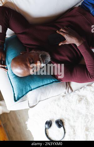 Vue en hauteur d'un homme âgé d'amérique africaine chauve avec un téléphone portable dormant sur un canapé dans une cabine en rondins Banque D'Images