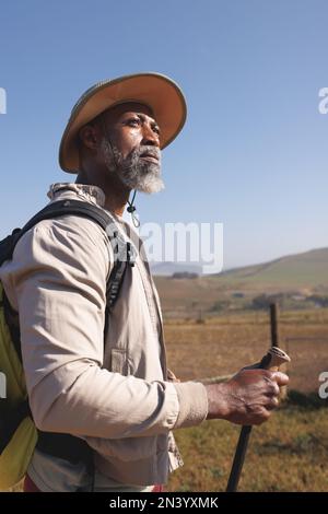 Homme senior afro-américain barbu portant un chapeau avec des bâtons de randonnée debout contre un ciel bleu clair Banque D'Images