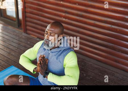 Vue à grand angle de l'homme senior chauve d'amérique africaine méditant dans la pose de prière sur le tapis à la cabane en rondins Banque D'Images