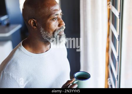 Homme senior chauve afro-américain attentionné avec tasse de café regardant par la fenêtre dans la cabane en rondins Banque D'Images