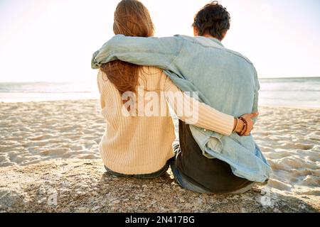 Mon idée de paradis est d'être avec vous. Vue arrière d'un jeune couple aimant assis les uns à côté des autres sur un rocher à la plage. Banque D'Images