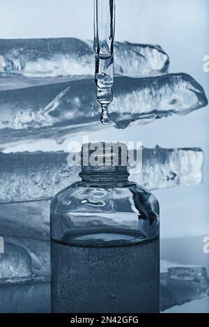 Une bouteille de sérum facial et une pipette avec une goutte sur le fond blanc avec des morceaux de glace fondue. Banque D'Images