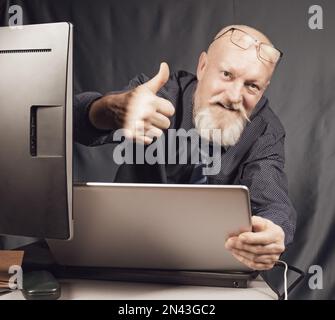 Travail au bureau, un homme âgé gai avec une barbe grise devant un ordinateur portable est satisfait du résultat. Banque D'Images