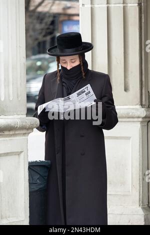 Le jour d'hiver, un juif hassidique lit un dépliant devant une synagogue Satmar avec une écharpe attachée sur sa bouche. À Williamsburg, Brooklyn, New York. Banque D'Images