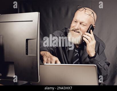 Travail de bureau - homme âgé gai avec une barbe grise parlant au téléphone tout en étant assis devant un ordinateur portable. Banque D'Images
