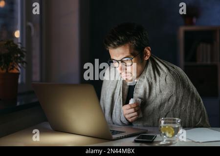 Jeune homme souffrant de la grippe et du mauvais rhume, travaillant à distance en auto-isolement avec un ordinateur portable tard dans la soirée de la maison, regardant les nouvelles sur le coronavirus, à la recherche d'un remède. Social distance, rester à la maison Banque D'Images