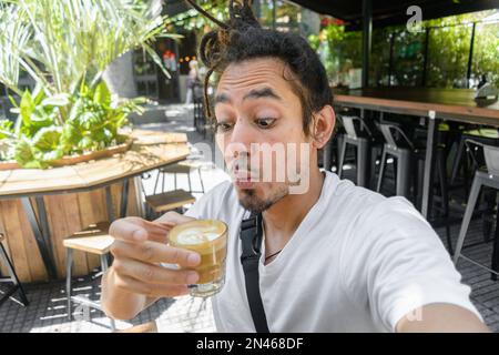 Vénézuélien latino jeune homme avec des dreadlocks prenant un portrait de selfie debout dans un restaurant à Buenos Aires soufflant un café parce qu'il est très chaud, Banque D'Images