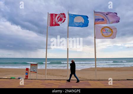 Villajoyosa Costa Blanca Espagne 02.08.2023 Plage Pavillon bleu. Homme marchant le long de la promenade en bord de mer carrelée en hiver. Trois poteaux avec banderoles fleuries Banque D'Images