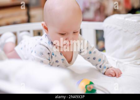 Le nouveau-né se leva sur les poignées dans un berceau avec un drap blanc. L'enfant s'intéresse aux premiers jouets éducatifs. Banque D'Images