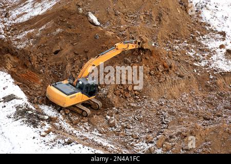 La pelle sur chenilles ramasse la terre avec un godet, vue de dessus. Travaux de terrassement en hiver, excavation sur un chantier de construction Banque D'Images