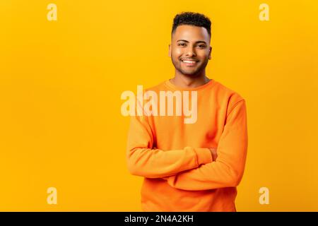 Portrait d'un jeune indien charmant et satisfait, décontracté, debout avec des bras croisés et regardant l'appareil photo avec un sourire éclatant. Studio d'intérieur tourné sur fond orange Banque D'Images