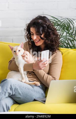 Souriant redacteur publicitaire tenant le téléphone cellulaire et chat oriental près de l'ordinateur portable sur le canapé, image de stock Banque D'Images
