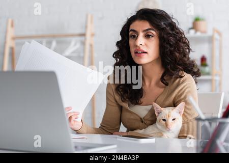 Brunette copywriter tenant des papiers et chat oriental près de l'ordinateur portable à la maison, image de stock Banque D'Images