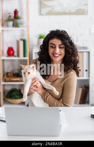 Souriant redacteur publicitaire tenant chat oriental près d'ordinateur portable à la maison, image de stock Banque D'Images