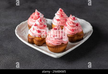 Petits gâteaux faits maison décorés de crème et de saupoudres en forme de coeur pour la Saint-Valentin dans une assiette en forme de coeur sur fond gris Banque D'Images