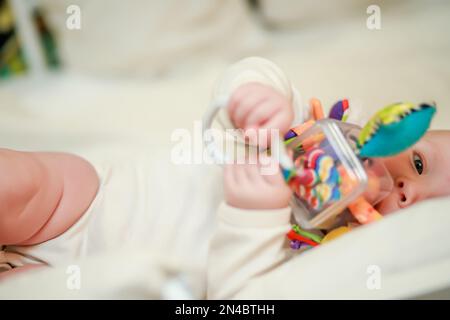 Un nouveau-né se trouve à côté et tient un jouet éducatif entre les mains. Jouet pour les sensations tactiles et l'étude des formes et des couleurs des tout-petits. Defocuse Banque D'Images