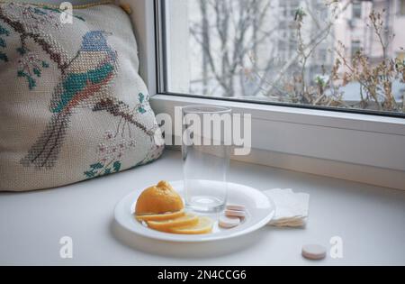 Froid de saison: Une tasse en verre vide, citron en tranches, pilules instantanées et vaporisateur nasal avec serviettes en papier sur le rebord de la fenêtre. À côté du modèle vintage brodé p Banque D'Images