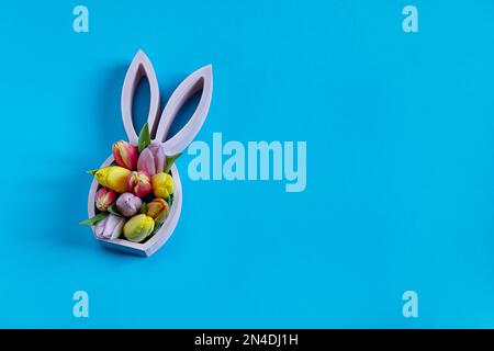 Figurine de lapin de pâques en bois avec tulipes de printemps sur fond bleu, concept minimal de pâques, plat avec espace de copie Banque D'Images