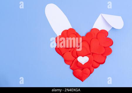 Carte de vœux créative. Imitation du lapin de Pâques. Un cœur rouge et des oreilles de papier blanc sur fond bleu. Le concept minimum de vacances de printemps. Banque D'Images