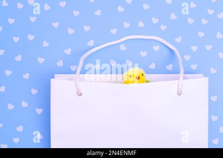 Sac à provisions en papier blanc avec poulet moelleux jaune sur fond bleu avec coeurs. Shopping pour Pâques. Concept cadeaux de Pâques, préparation, vacances su Banque D'Images