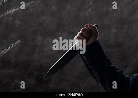 Une personne en chemise noire à manches longues tenant un couteau contre un fond gris Banque D'Images