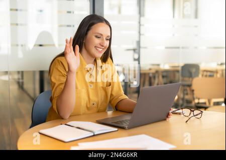 Concept de réunion vidéo. Une jeune femme joyeuse a un appel vidéo sur l'ordinateur portable au bureau, en agitant le salut dans la webcam, saluant les interlocuteurs en ligne, impliqué en ligne réunion vidéo avec l'équipe de travail Banque D'Images