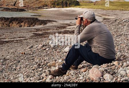 Man 60+ assis sur une plage de pierres Camas Cuil et t-Saimh, sur le côté ouest d'Iona, en Écosse, en regardant la mer à travers des jumelles Banque D'Images