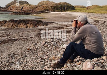 Man 60+ assis sur une plage de pierres Camas Cuil et t-Saimh, sur le côté ouest d'Iona, en Écosse, en regardant la mer à travers des jumelles Banque D'Images