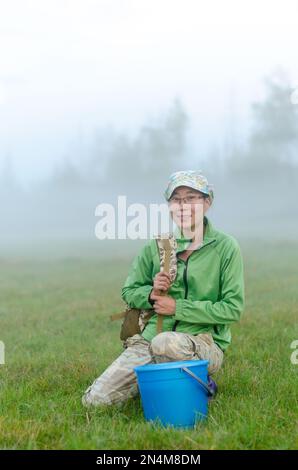 Yakut asiatique fille dans un chapeau et des lunettes assis souriant et tenant la sangle d'un sac à dos au seau avant de cueillir des baies sauvages dans le brouillard blanc b Banque D'Images